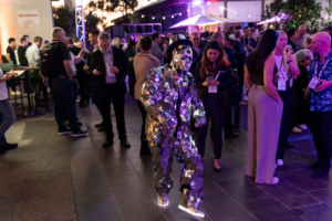 A street performer dressed in a mirrored suite walking through a crowd of people who are drinking and talking at an event at night.